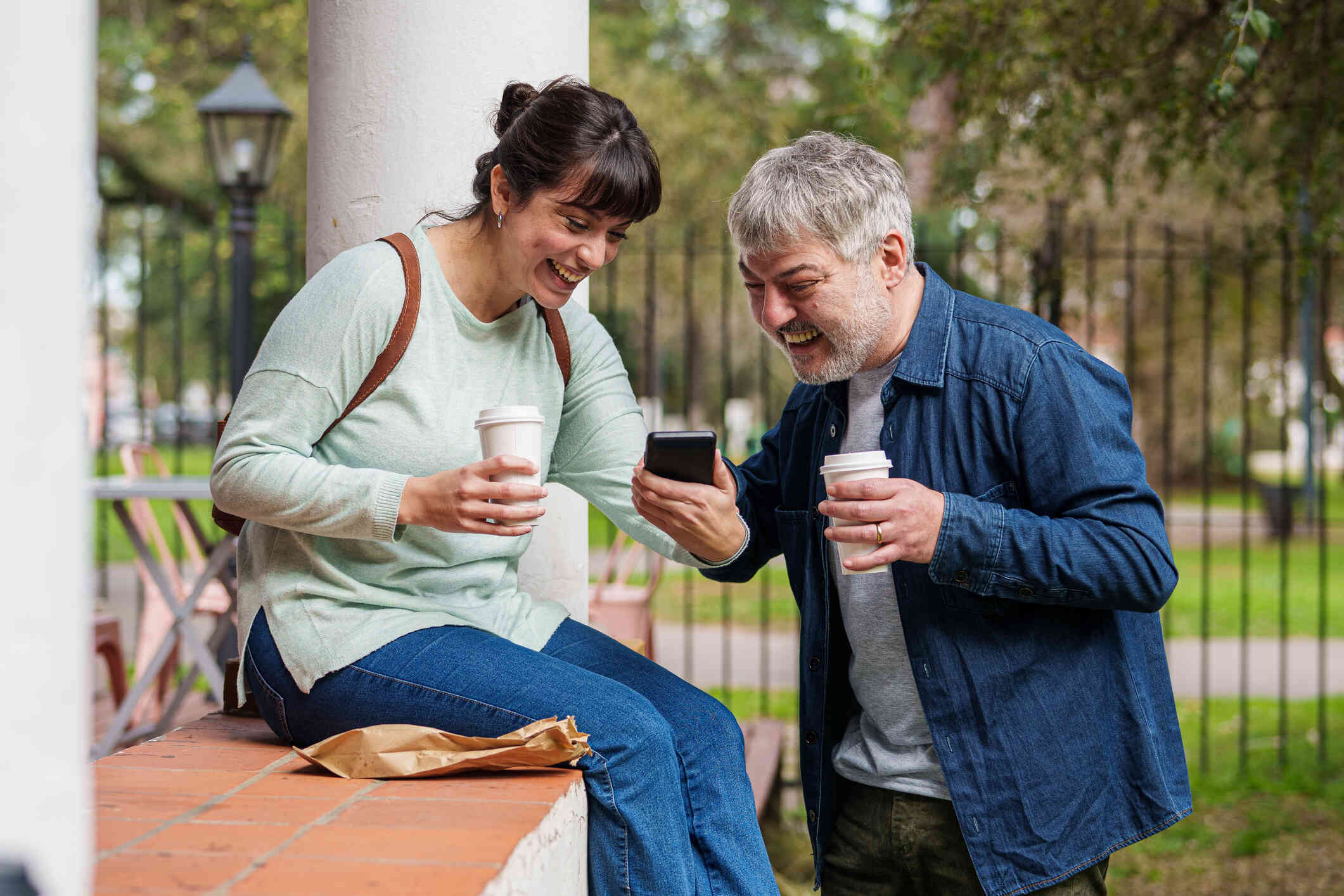 A woman in a greens shirt sits on a ledge while holding a top-go cup and shows the man infront of her her phone as they both smile.